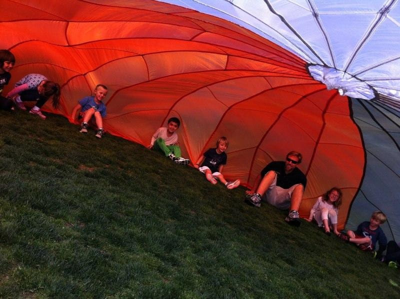7 students and teacher sitting inside a parachute on a grass field.