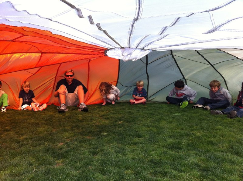 7 students with teacher sitting on a grass field under a very large parachute