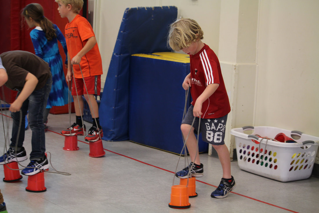 4 young students using cup stilts with ropes to hold practicing balance walking around the gym