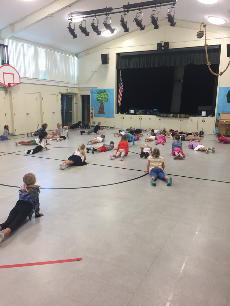 Students performing cobra yoga pose in a gym with a parent volunteer leader
