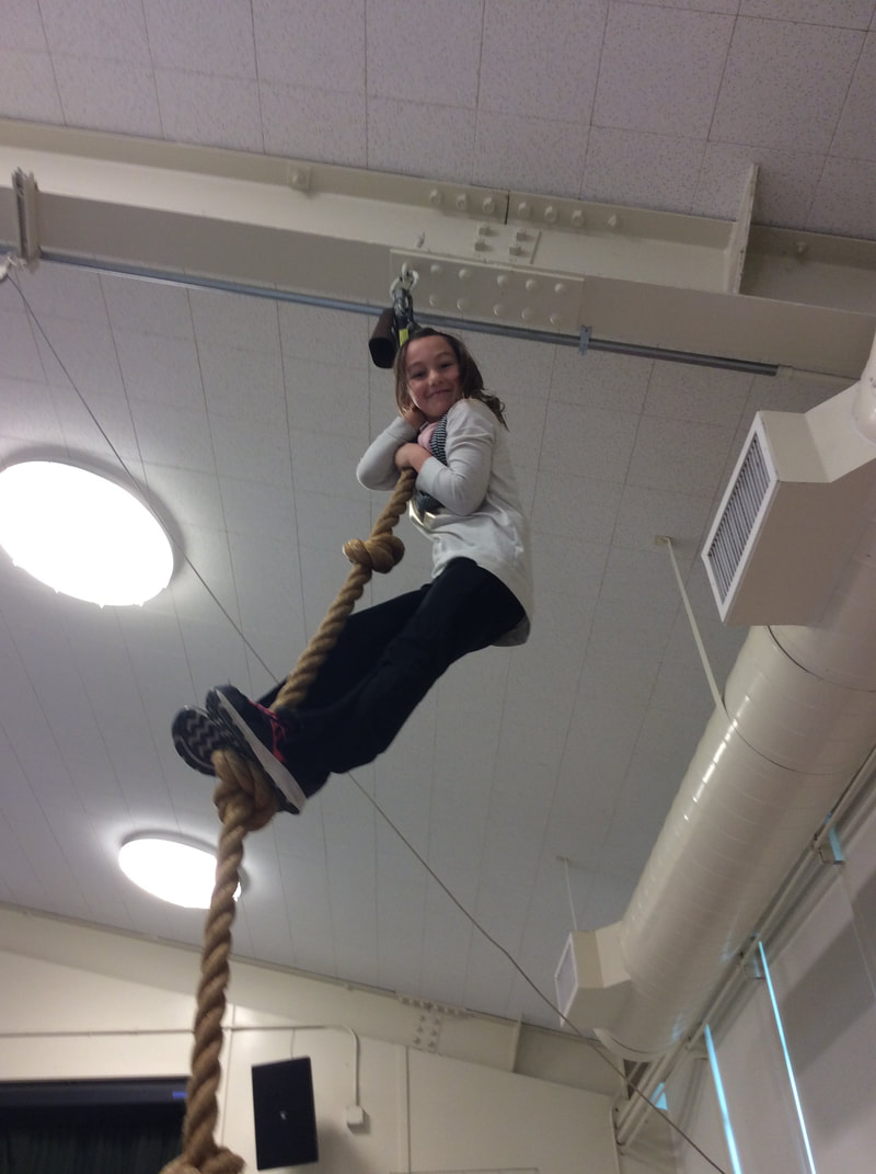 Young girls smiling at the top of the rope that extends up to the ceiling in the gym.